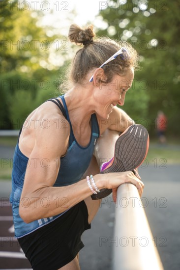 Woman smiling as she stretches her leg on a running track surrounded by summer nature, middle distance runner Katharina Jaiser, Gechingen, Germany