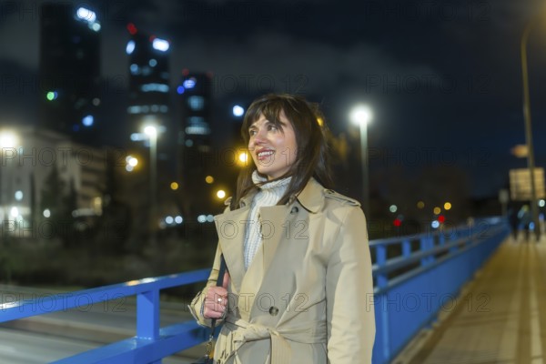 Woman wearing a trench coat and warm sweater standing on a bridge, looking at the illuminated city skyline with modern skyscrapers and night lights in an urban setting