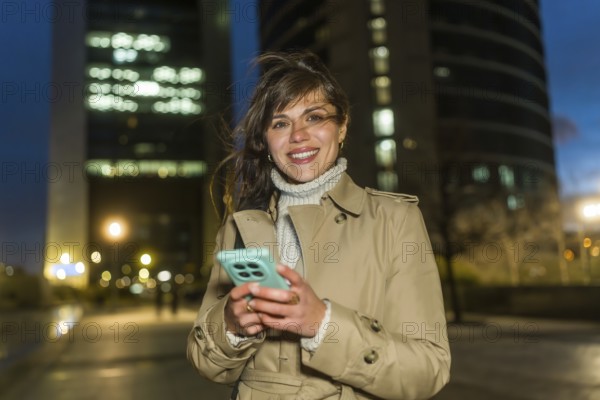 Young woman in a trench coat stands on a lit city street at dusk, smiling at the camera while holding a smartphone, conveying modern connectivity and urban lifestyle