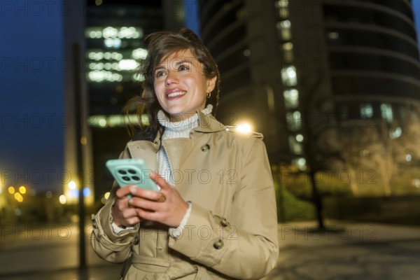 Woman smiling, holding a smartphone, and looking away in an illuminated urban environment with modern buildings in the background during the evening, representing communication and urban lifestyle