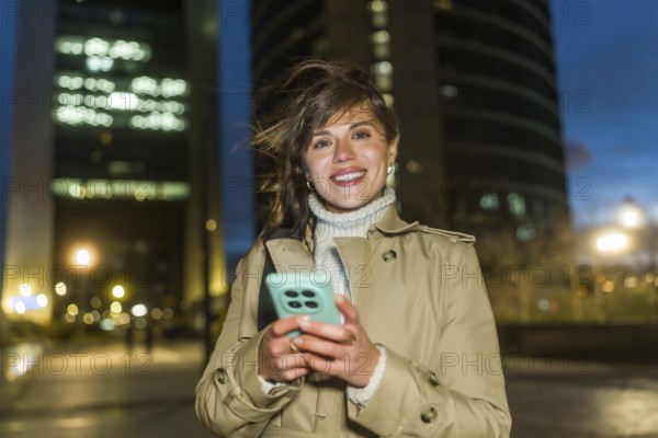 Young woman smiling, holding her smartphone, and looking at the camera outdoors in a modern city setting at dusk, with office buildings and bokeh lights in the background