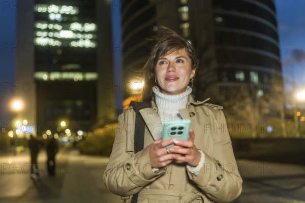 Woman standing outdoors at night, using a smartphone for communication and networking, dressed in a trench coat with city lights and blurred buildings creating an urban background