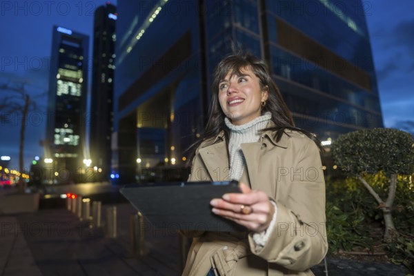 Woman stands at twilight in a glowing downtown, smiling while using a tablet, blurred skyscrapers and lit streets suggest urban tech lifestyle, business and connectivity