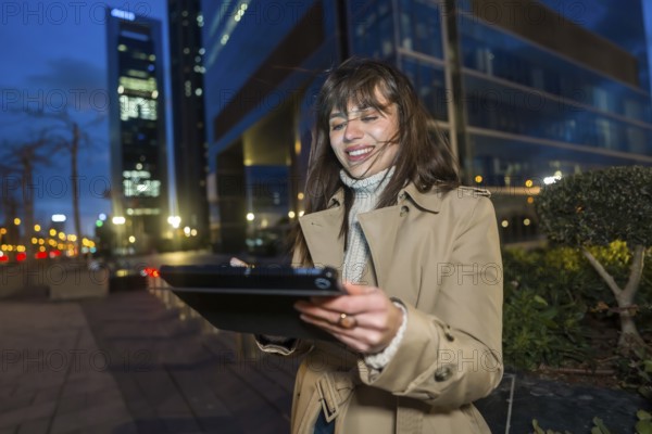 Young professional woman in a trench coat standing in a lit downtown at night, smiling while using a tablet, enjoying urban life and wireless connection among skyscrapers