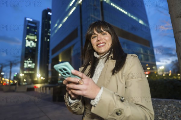 Young professional woman smiling while using her smartphone for networking and social media in a modern urban business district at dusk, confident and connected outdoors