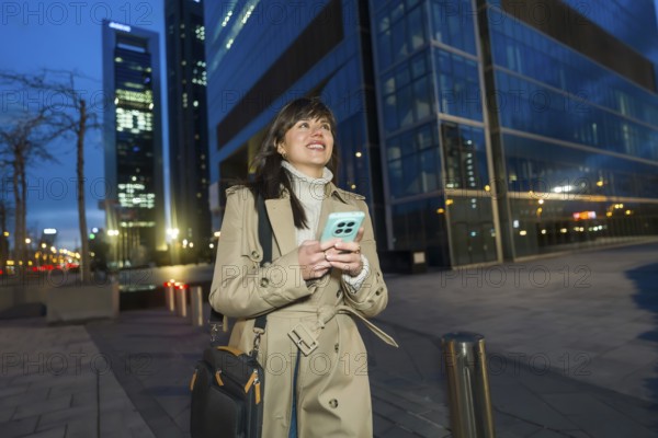 Young woman walks confidently along a modern city street at dusk, trench coat and turtleneck, holding smartphone and smiling as she connects online amid skyscrapers and evening lights
