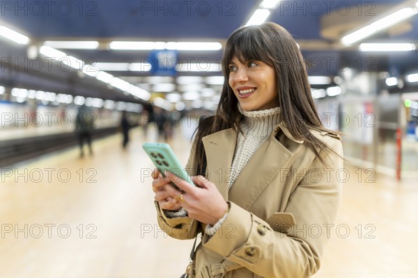 Woman standing on a train station platform, smiling and looking away while sending messages or browsing on her smartphone, representing urban connectivity and modern commuting