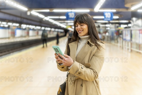Young woman standing on a subway platform and smiling while checking her mobile phone, actively engaged with her device as she waits for public transport