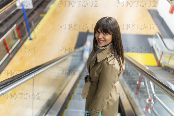 Woman in a trench coat smiling confidently while riding down an escalator in a modern subway station, representing urban lifestyle, daily commute, and public transportation