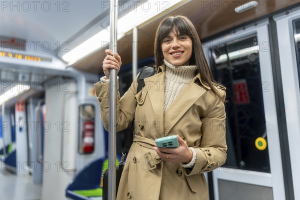 Young smiling woman in a trench coat stands in a subway car, holding her smartphone while commuting through the city, scrolling and enjoying her urban journey