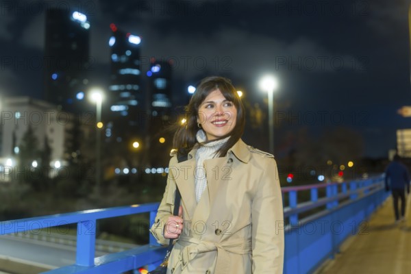Woman in trench coat on an urban bridge at night, smiling and looking away as illuminated skyscrapers and city lights of madrids cuatro torres sparkle in the evening haze