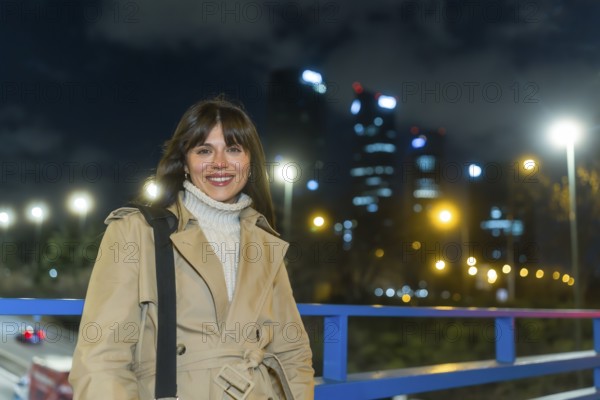 Young woman wearing a trench coat and turtleneck sweater smiling confidentially, standing outdoors on an urban bridge overlooking modern buildings and city lights at night