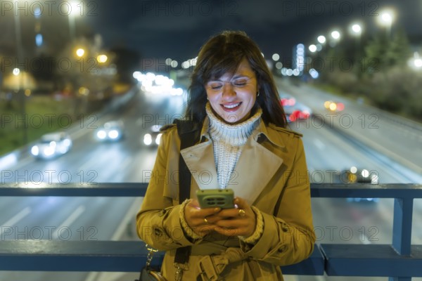 Young woman smiling and engaging with a mobile phone, standing on an urban bridge overlooking a busy highway with bokeh lights at night, embodying connectivity and modern communication