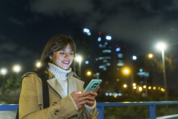 Young woman smiling as she types on her smartphone on a city street at night, illuminated by bokeh lights and blurred urban buildings, casually dressed and focused on her screen