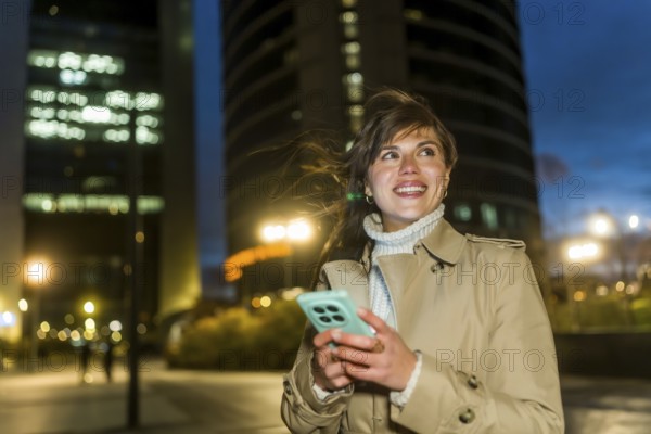 Young woman smiling, looking up while navigating her smartphone, dressed in a trench coat and turtleneck, enjoying a pleasant stroll through a modern city district during the evening