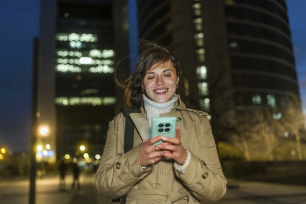 Young woman smiling while walking through the city at night, engaging with her mobile phone and communicating digitally in an urban environment filled with illuminated buildings