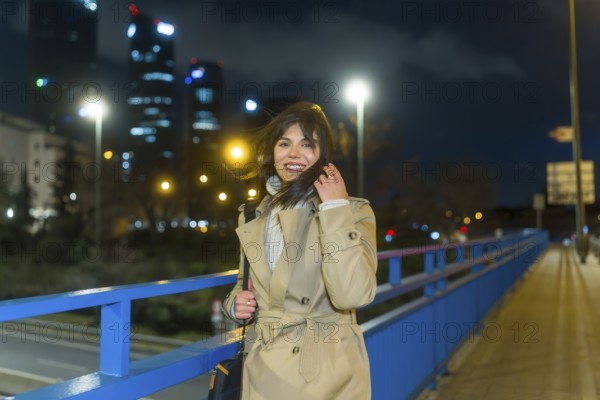 Young woman in a trench coat smiling, touching her hair, and looking at the camera while standing on a bridge in an urban city at night with blurred skyscrapers and street lights in the background