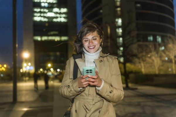 Young woman in a trench coat smiling, walking through a city at night, connecting with her smartphone, experiencing urban lifestyle and happy communication under city lights