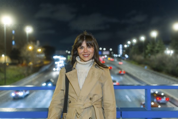 Woman smiling and standing on a bridge over a blurred highway with car lights creating bokeh, reflecting urban movement, modern city life, and evening commute