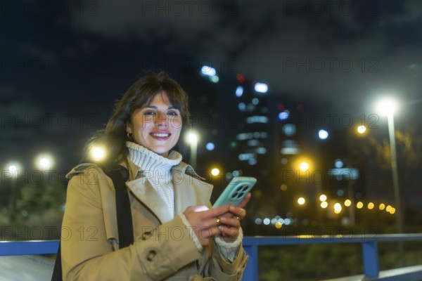 Woman standing on a city bridge at night, smiling and typing on her mobile phone, with illuminated skyscrapers and streetlights creating a bokeh background