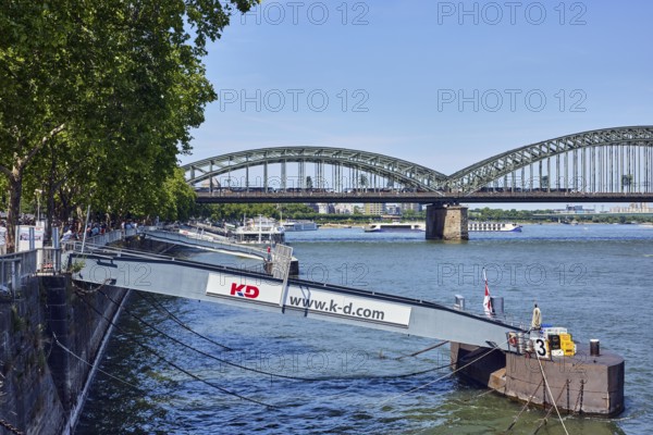 Rhine river, pedestrian and railway bridge Hohenzollern bridge, steel arches, general architecture, pier, gangway, Cologne-Düsseldorfer Deutsche Rheinschiffahrt GmbH, quay wall, water surface with small waves, trees, blue sky, cloudless, Leystapelwerft, Cologne, district-free city, North Rhine-Westphalia, Germany