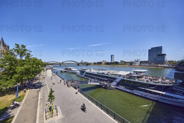 Rhine river, waterfront, sightseeing boats, general architecture, high-rise buildings, modern architecture, pedestrian and railway bridge Hohenzollern Bridge, trees, cyclists, pedestrians and visitors to a sightseeing boat as a side theme, elevated perspective, blue sky, cirrus clouds, Deutz bridge, Leystapelwerft, Cologne, district-free city, North Rhine-Westphalia, Germany