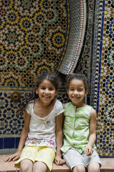 Two smiling girls in front of a wall with colorful tiles, alley in the old town, Souk, Fez El Bali, Medina, UNESCO World Heritage Site, Fez, Morocco
