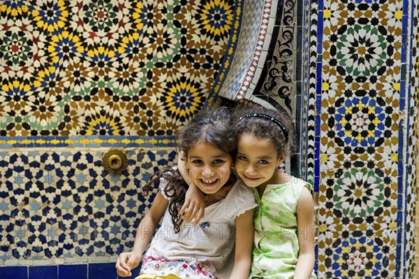 Two smiling girls in front of a wall with colorful tiles, alley in the old town, Souk, Fez El Bali, Medina, UNESCO World Heritage Site, Fez, Morocco