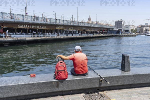 Istanbul, Turkey. June 23rd 2025. A mam sits fishing on the dockside in Eminonu beside the famous Galata Bridge with its fish restaurants and the iconic Galata Tower, Istanbul, Turkey
