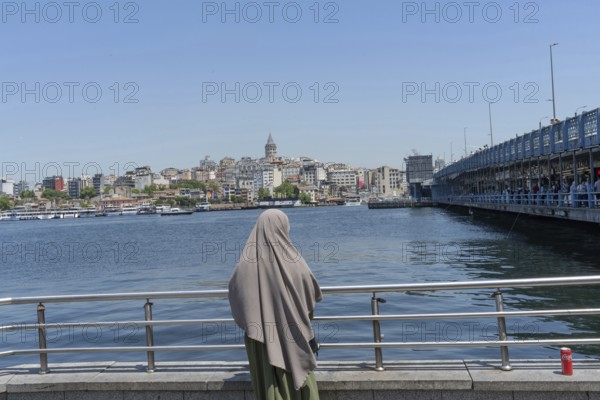 Istanbul, Turkey. June 23rd 2025. A Muslim women looks out across the Golden Horn towards the Galata Tower an iconic landmark of the largest city in Turkey