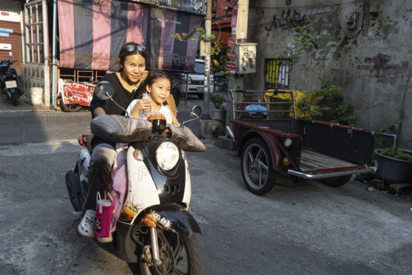 Bangkok, Thailand. March 18th 2025. A mother with her daughter rides a motor scooter through the narrow streets of Talat Noi, Bangkok, Thailand, Asia