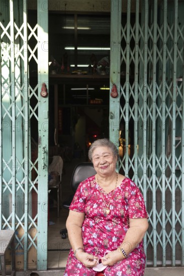 Bangkok, Thailand. March 18th 2025. A smiling older lady sitting outside a shop house in the streets of Talat Noi, Bangkok, Thailand, Asia
