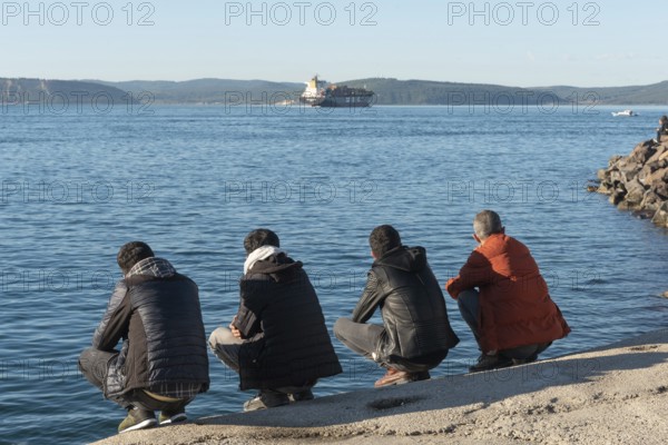 Canakkale, Turkey. February 18th 2022 Turkish men squatting beside the Dardanelles Straight opposite the Gallipoli Peninsular, the harbour of the port city of Canakkale, Western Turkey