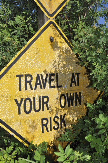 A weathered yellow road sign with the text 'Travel at Your Own Risk, 'flanked by leaves, Grand Staircase Escalante National Monument, Utah, USA