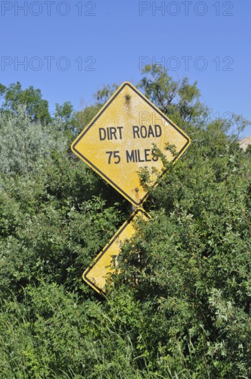 A yellow road sign with the words 'Dirt Road 75 Miles' appears from lush vegetation, Grand Staircase Escalante National Monument, Utah, USA