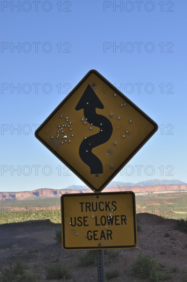 A road sign with serpentines strewn with bullet holes in a bare landscape with rolling hills in the background, Grand Staircase Escalante National Monument, Utah, USA