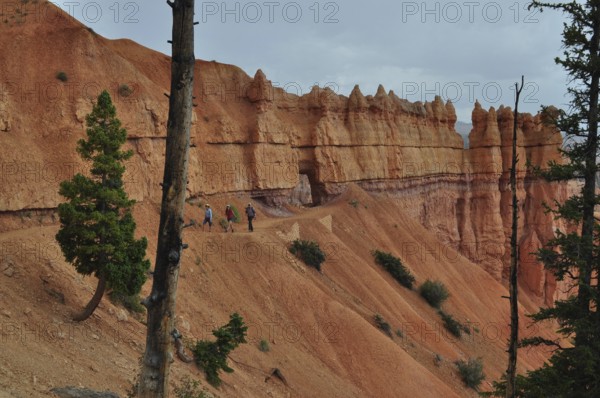 Hikers on a trail along dramatic red cliffs under looming clouds, Bryce Canyon National Park, Utah, USA