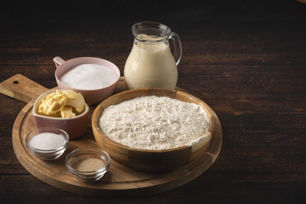 Baking ingredients on a wooden table with a bowl of flour, sugar, and a pot of milk