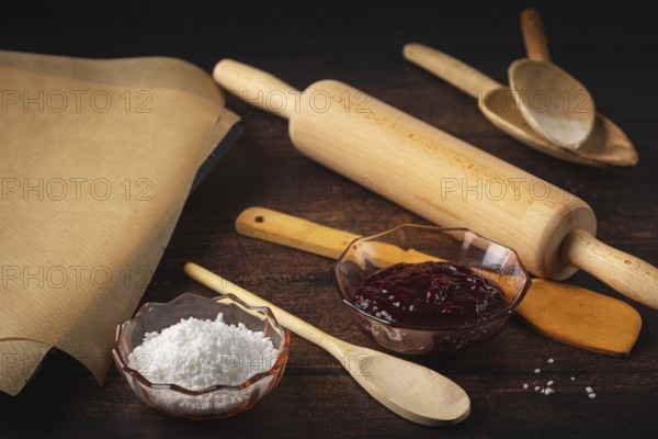 Rolling pin, cooking spoon and baking paper next to jam and sugar on a dark wooden table in a rustic atmosphere