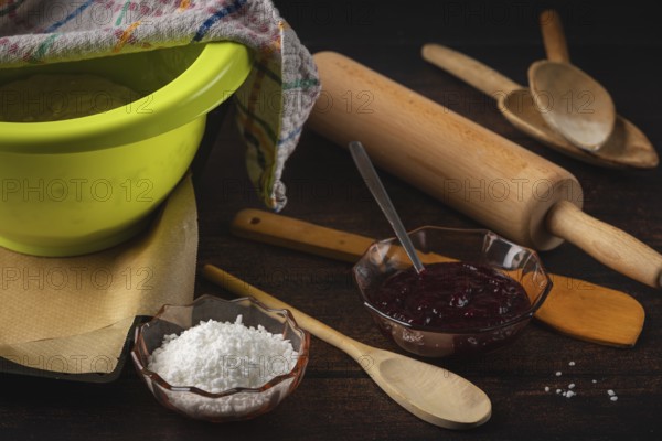 Dough bowl next to rolling pin, cooking spoon, baking paper, jam and sugar on a dark wooden table