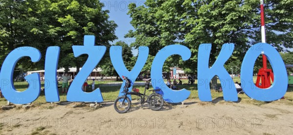 A bicycle stands in front of a large blue lettering of 'Gizycko' on a sandy beach, Masuria bicycle tour, Poland