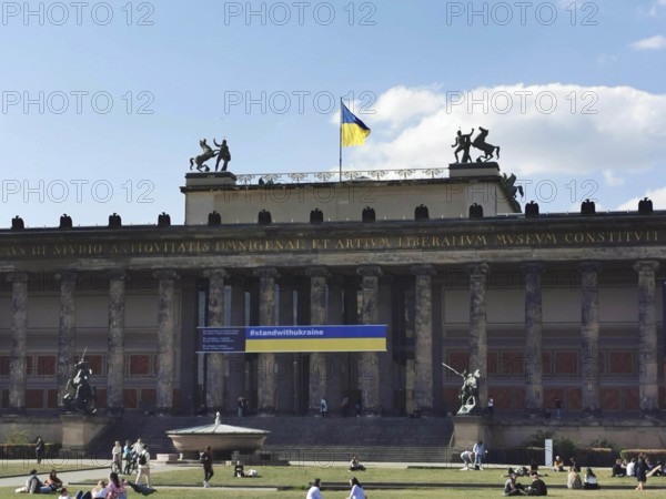 People relax on the lawn in front of the old museum on Museum Island in Berlin where the Ukrainian flag is blowing in the wind, Berlin