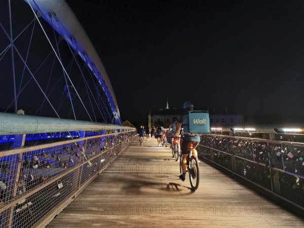 Delivery service Cyclists cross an illuminated bridge with railings on which countless padlocks hang with an urban background, Krakow, Poland