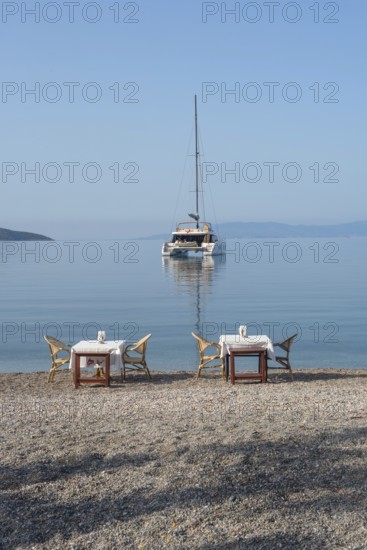 Bodrum, Mugla, Turkey. April 22nd 2022 Restaurant tables on the beach with a Catamaran moored in Bodrum harbour, a popular sailing and cruise destination along the Aegean coast in south west Turkey
