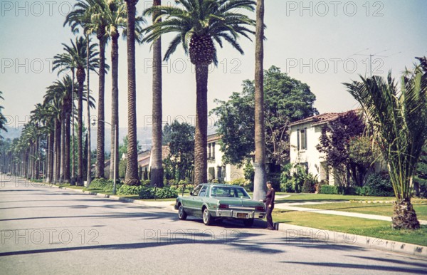 Woman standing by Chrysler Plymouth Volarét car in Beverly Hills, California, characterized by its signature towering palm trees, USA 1976