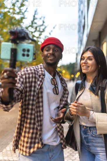 Diverse couple vlogging outdoor, enthusiastically recording video on a smartphone with a microphone and selfie stick, sharing experiences for social media content