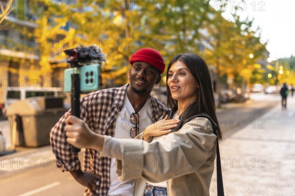 Diverse couple recording engaging video content on a smartphone with a gimbal stabilizer, producing media for social platforms while walking along a street during a clear autumn day