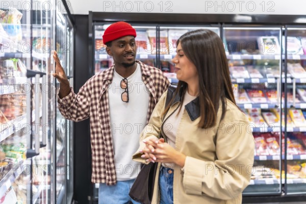 Diverse couple, a black man and a caucasian woman, making decisions about products from a refrigerated display while grocery shopping in a supermarket aisle