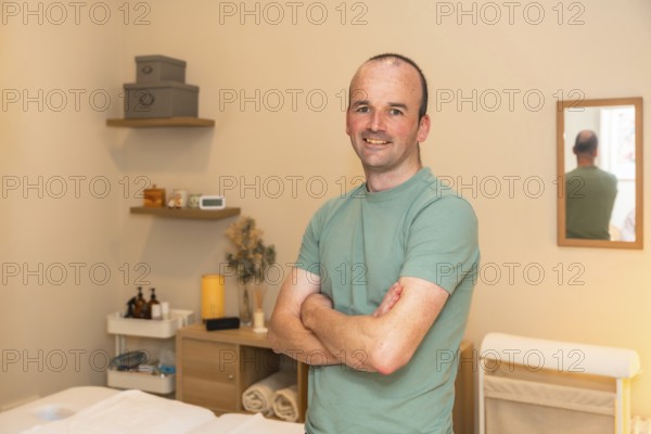 Professional man massage therapist smiling confidently, standing with crossed arms in a therapy room, ready to provide wellness and health services to clients