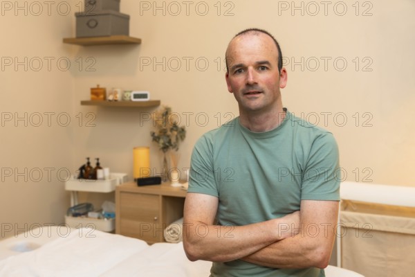 Male massage therapist owner standing with crossed arms in his wellness clinic, offering health, relaxation, and self care services in a professional spa environment